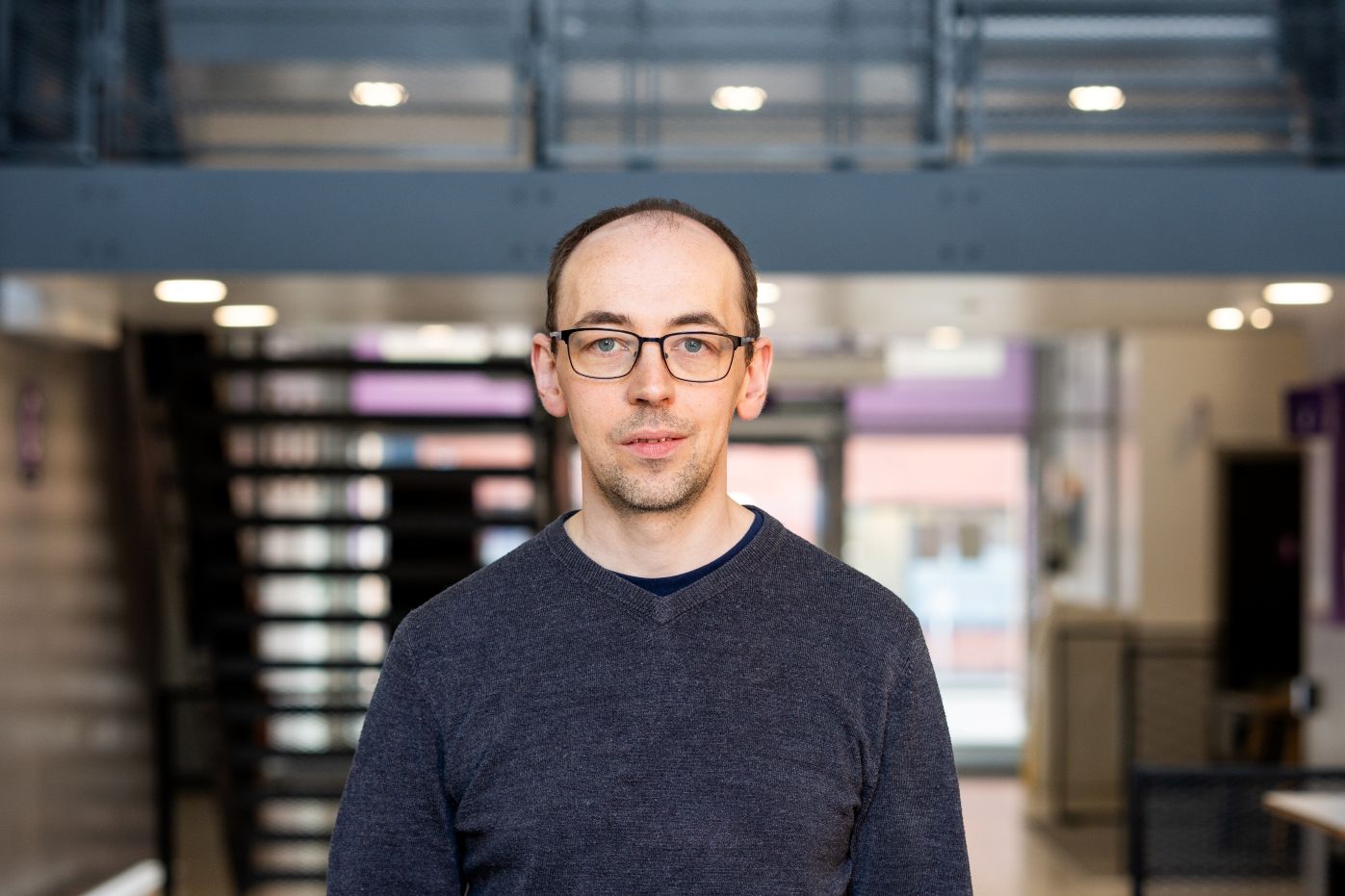 Photo of Stuart Christie, who is standing in the hallway wearing a dark grey sweater and glasses.